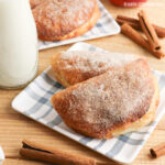 Two apple hand pies sitting on a blue plaid plate with cinnamon sticks and a glass bottle of milk in the background.