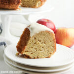 A close up view of a slice of apple bundt cake on a white plate with two red apples in the background.