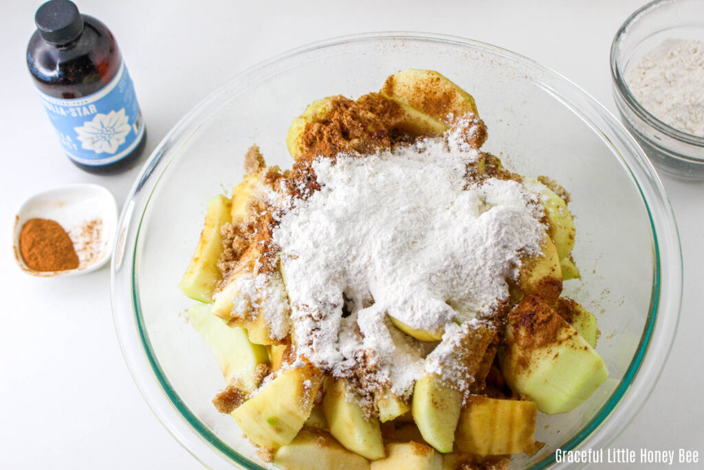 peeled and sliced apples in a clear mixing bowl topped with sugar, cinnamon and flour.