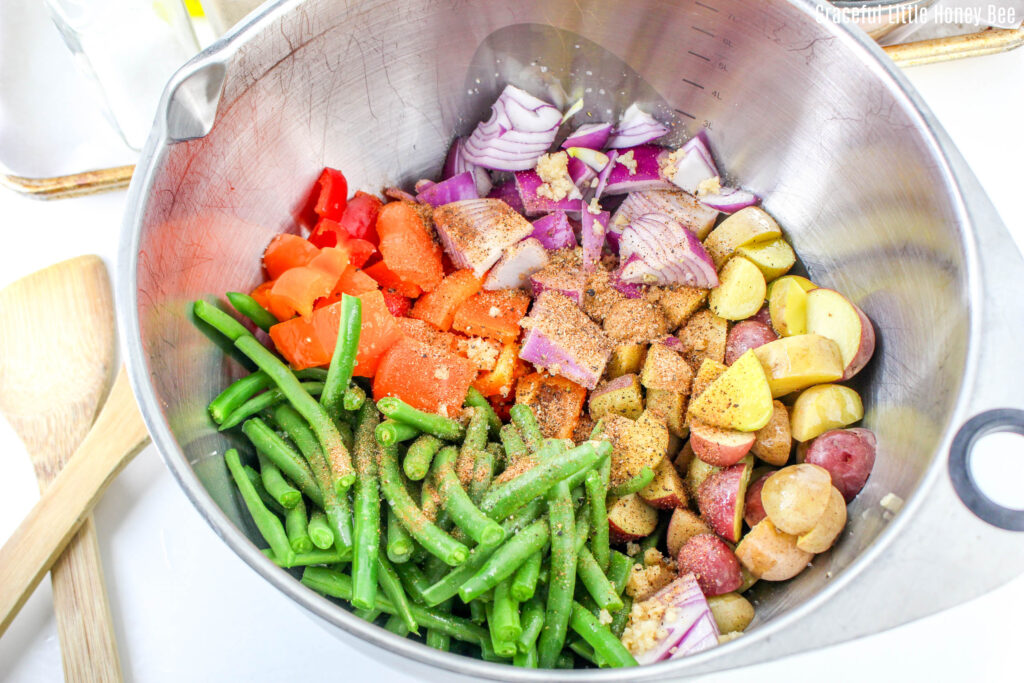 Metal mixing bowl filled with chopped vegetables and oil an seasoning before being mixed.