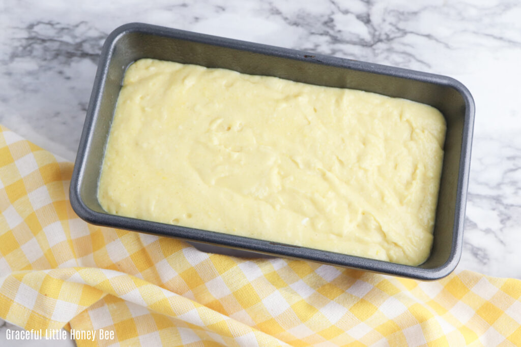 Lemon batter in a loaf pan with a yellow gingham tea towel.