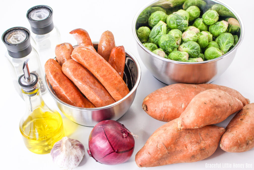 Sweet potatoes, a bowl of Brussels sprouts, a bowl of chicken sausage, a red onion, garlic, oil and salt and pepper sitting on a counter.