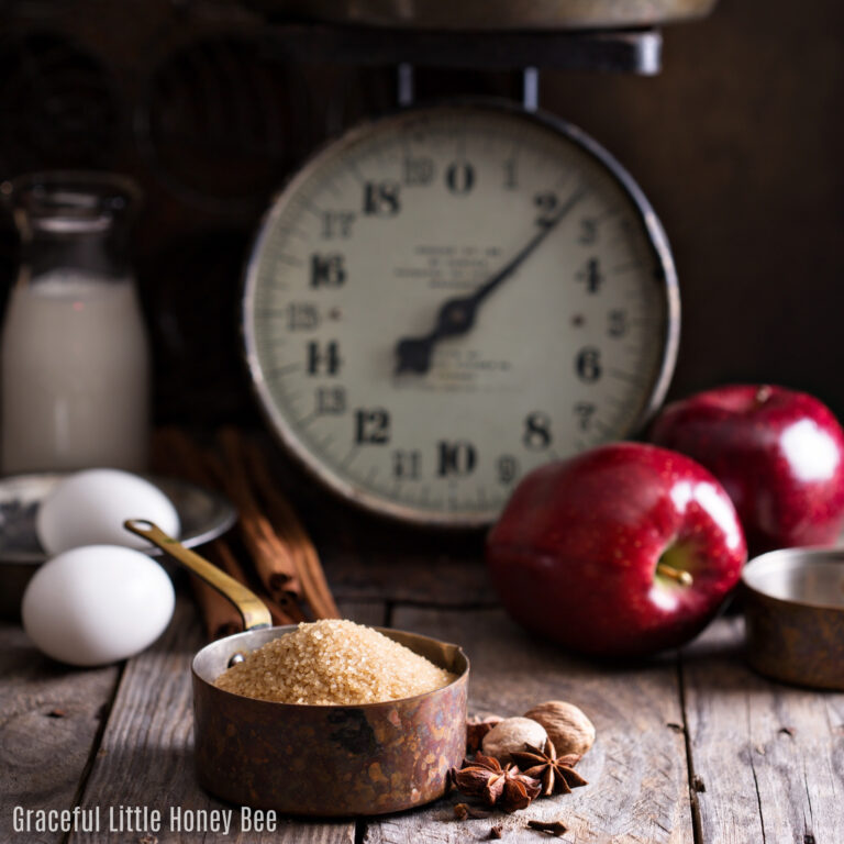 A kitchen scale, apples, eggs, milk, sugar and spices sitting on a counter.