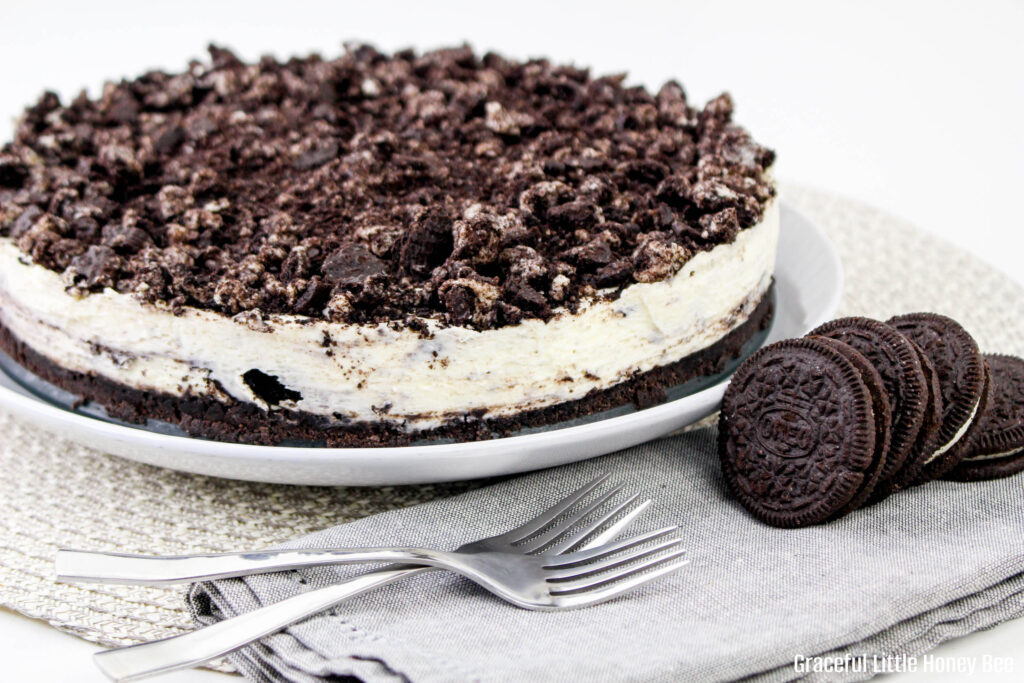 A whole Oreo cheesecake sitting on a white plate with Oreos and forks sitting next to it.