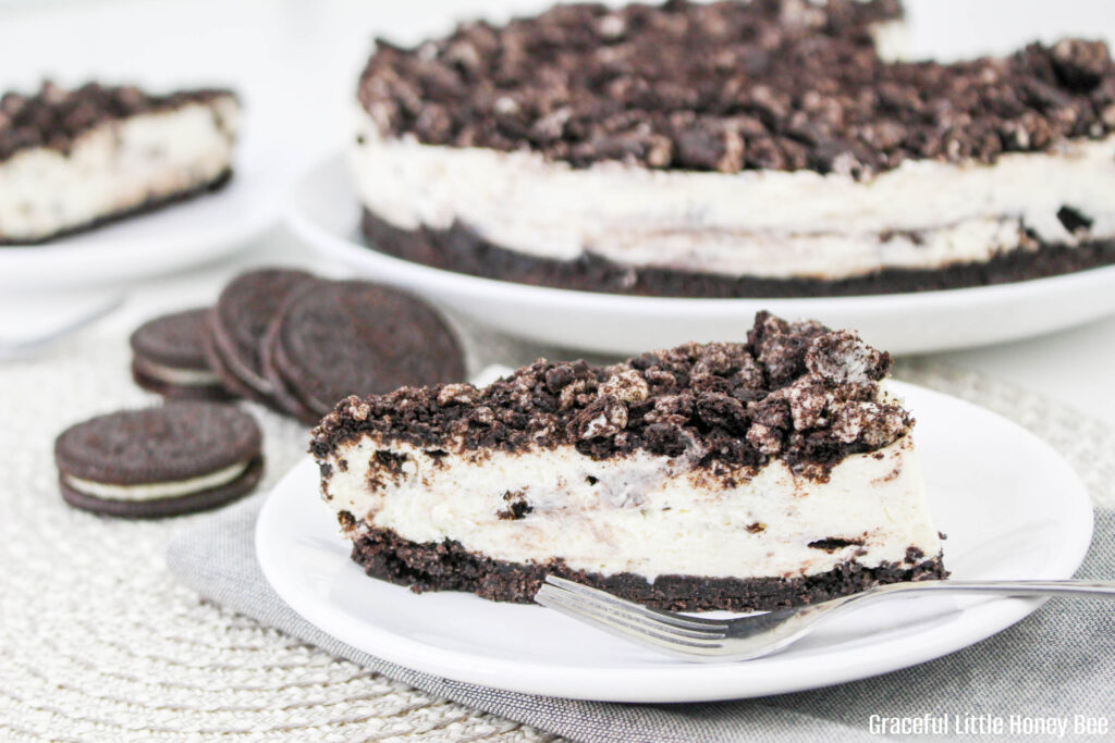 A slice of Oreo Cheesecake on a white plate with a fork and Oreos in the background.