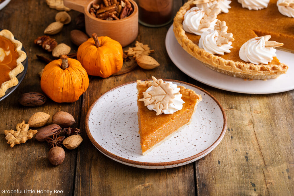 A slice of pumpkin pie topped with whipped cream sitting on a white plate with pumpkins in the background.