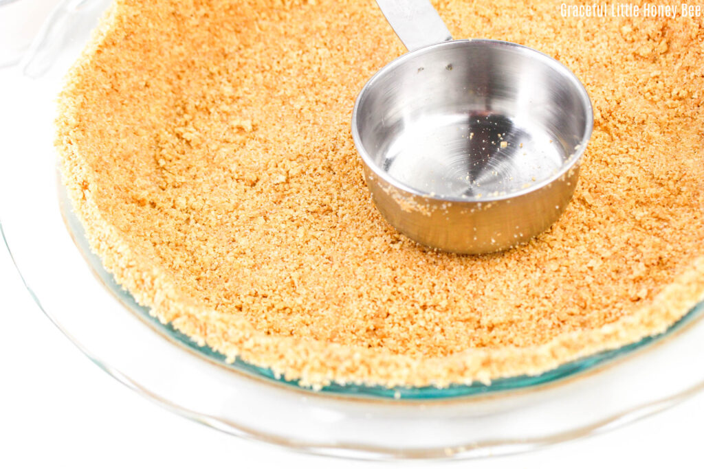 Graham cracker crust being pushed onto a pie plate with a measuring cup.