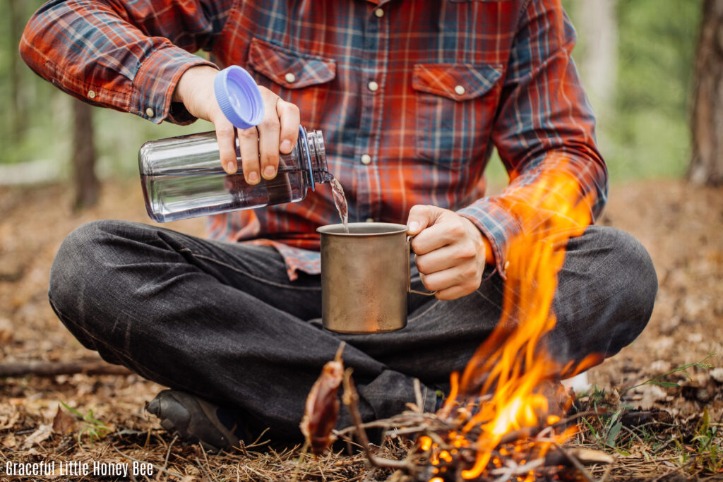 Man in plaid shirt pouring water into a coffee cup next to a campfire.