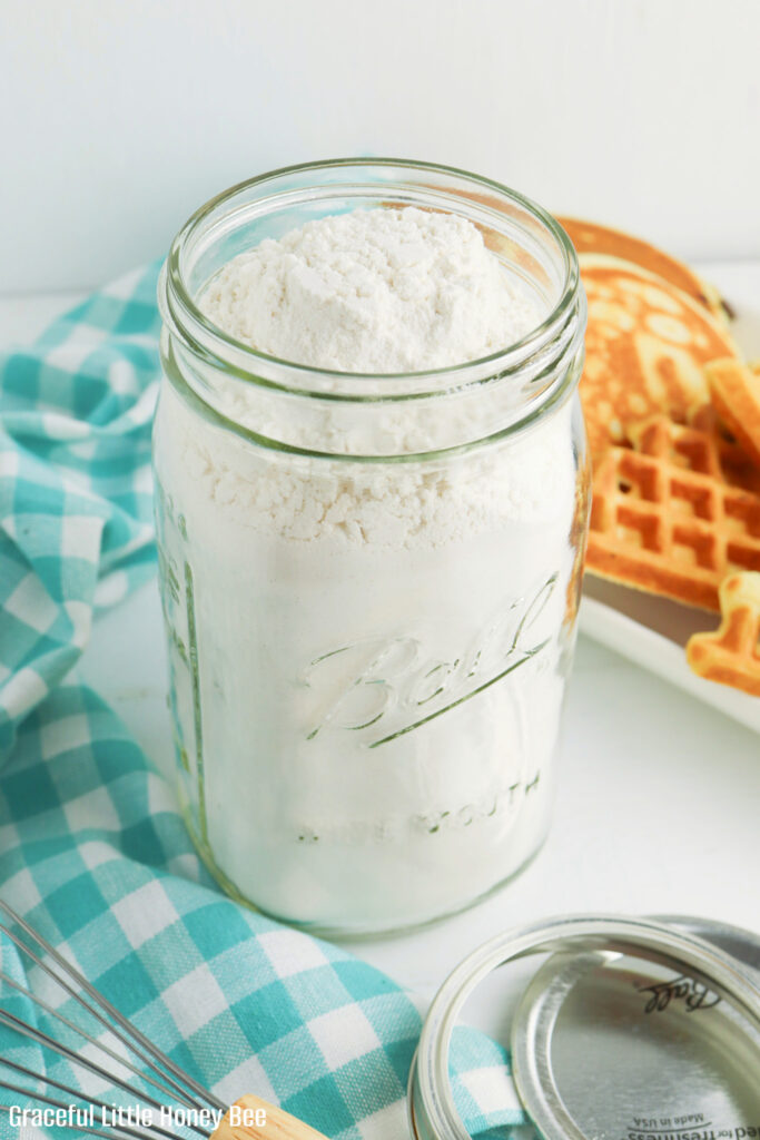 Pancake mix in a mason jar with a plate of pancakes and waffles in the background.