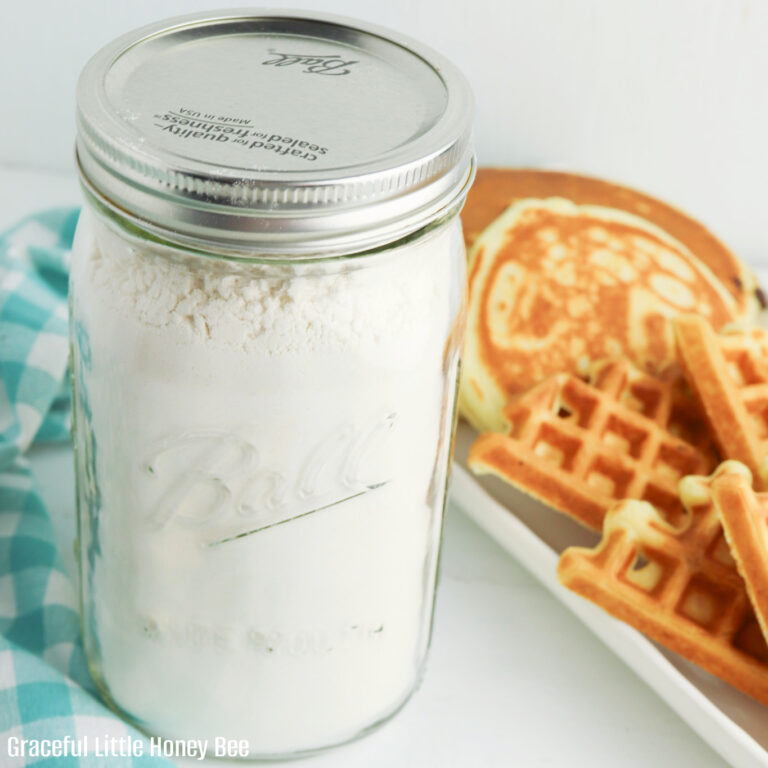 Pancake mix in a mason jar with a plate of pancakes and waffles in the background.