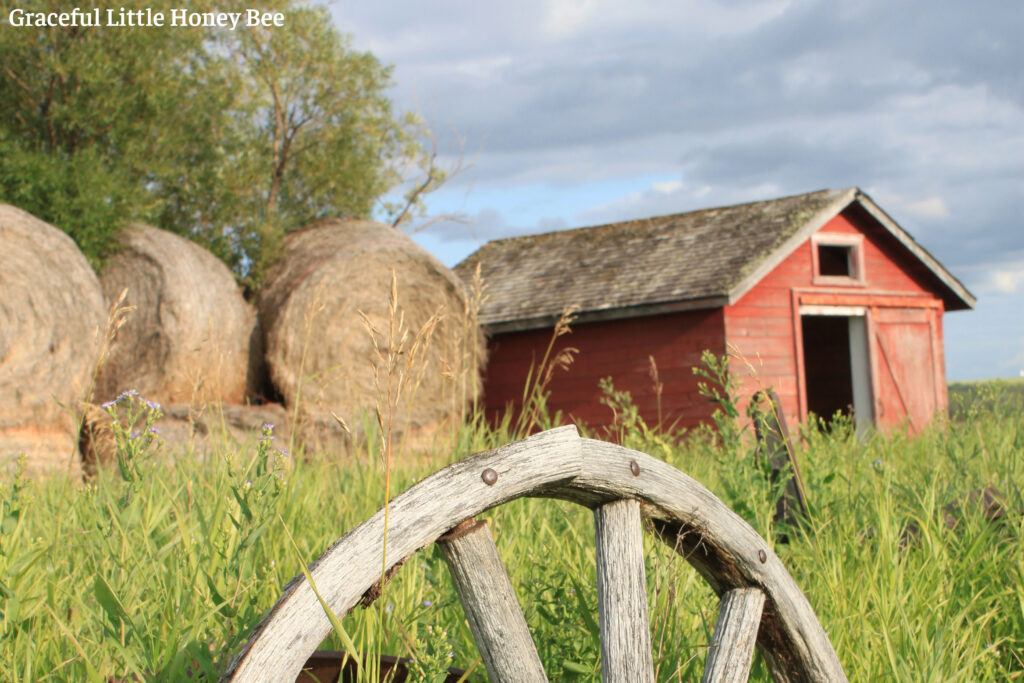 Red barn with hay bails and wooden wheel next to it.