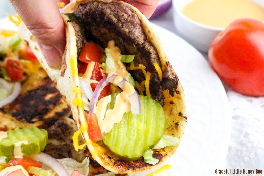 A close up view of someone holding up a smash burger taco topped with tomatoes, pickles and red onion.