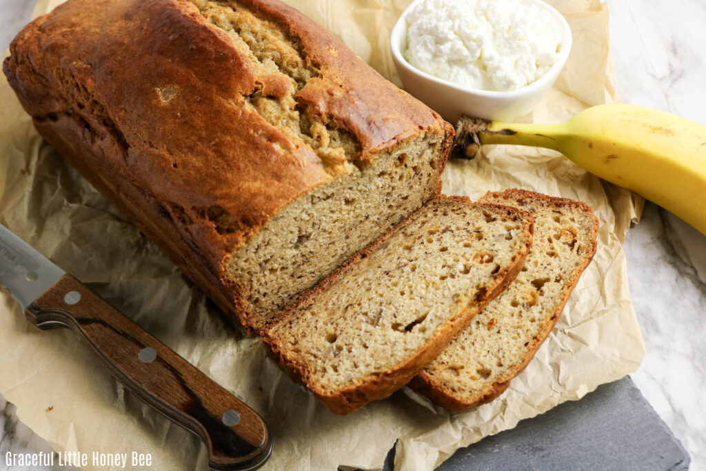 Loaf of banana bread with two slices cut sitting on a cutting board next to a bowl of cottage cheese and a banana.