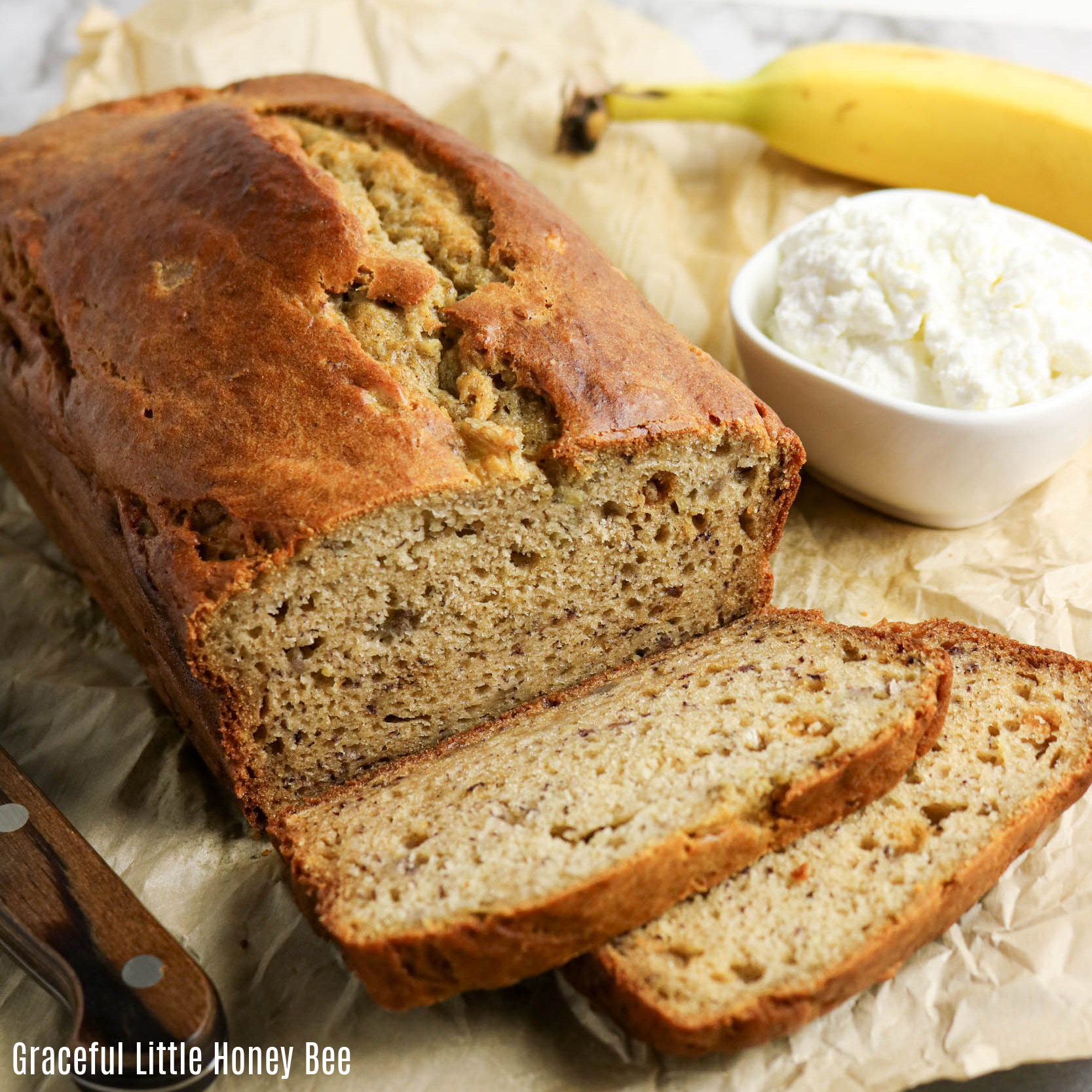 Loaf of banana bread with two slices cut sitting on a cutting board next to a bowl of cottage cheese and a banana.