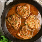 Four smothered pork chops in a cast iron pan with onion gravy.